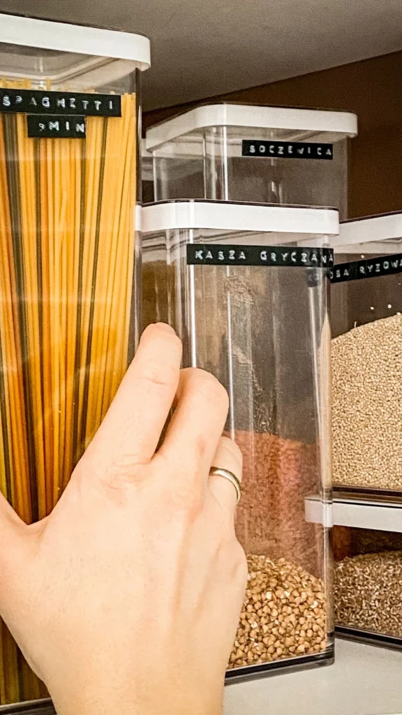 Ręka sięgająca po opisane pojemniki na produkty suche w uporządkowanej przestrzeni kuchennej.